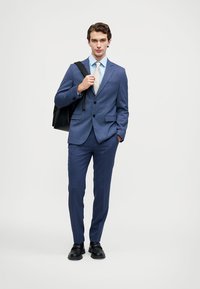 Sötétkék öltöny texturált felülettel, két gombos zakóval, hozzá illő szabott nadrággal, világoskék inggel és fekete cipővel. Fekete táska tartozik hozzá.