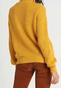 Pull en maille jaune moutarde avec un col haut, coupe ample, épaules tombantes et détails subtils sur le côté. Associé à un pantalon en velours côtelé marron.