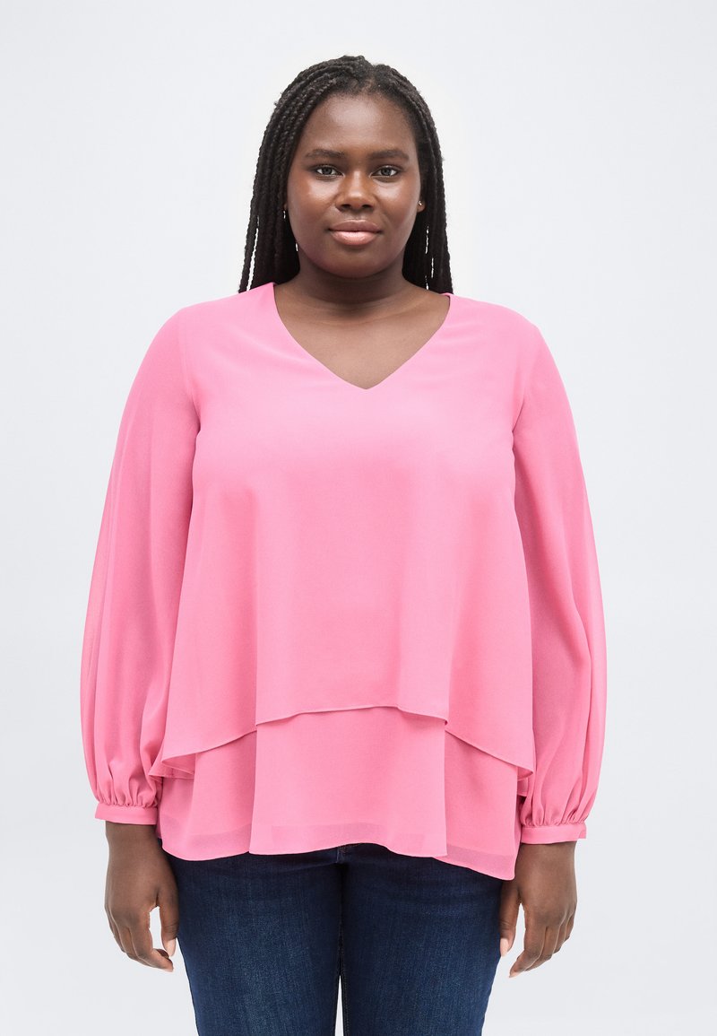 Γυναίκα με μακριά πλεξούδα, φορώντας ροζ μπλούζα με στρώσεις και σκούρο μπλε τζιν, στέκεται μπροστά σε απλό ανοιχτόχρωμο φόντο.