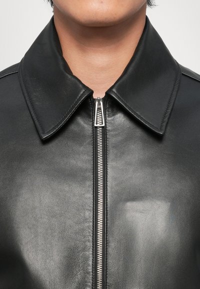 Zwarte leren jas met een prominente kraag, voorritssluiting en een gladde textuur. Het ontwerp heeft een klassieke stijl zonder zichtbare patronen.