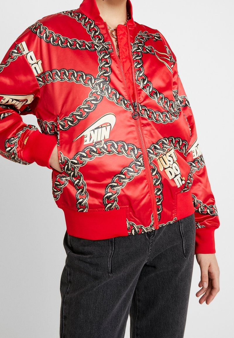 Röd satin bomberjacka med kedjemönster, guldfärgade detaljer och "Nike"-logotyp. Ribbad manschetter och fåll fulländar designen.