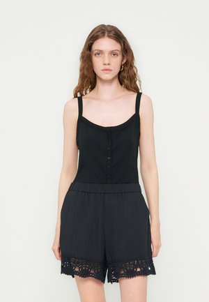 Młoda kobieta o długich falowanych włosach, ubrana w czarny bezrękawnik z guzikami i czarne szorty z koronkową lamówką, stojąca na jednolitym tle.