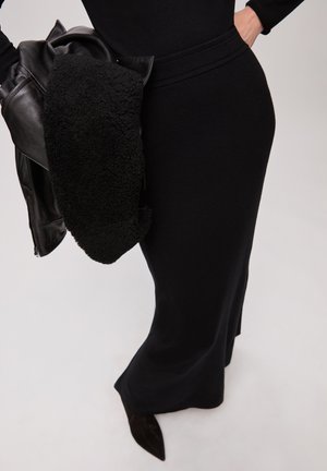 Femme portant une robe noire cintrée tenant une veste en cuir noire avec une doublure texturée sur le bras, sur un fond uni.
