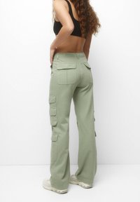 Kvinde iført lysegrønne cargobukser med flere lommer, kombineret med en sort crop top og hvide sneakers, stående mod en hvid baggrund.