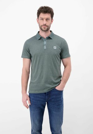 Mann trägt ein grünes gestreiftes Poloshirt und blaue Jeans, steht mit einer Hand in der Tasche vor einem einfarbigen weißen Hintergrund.