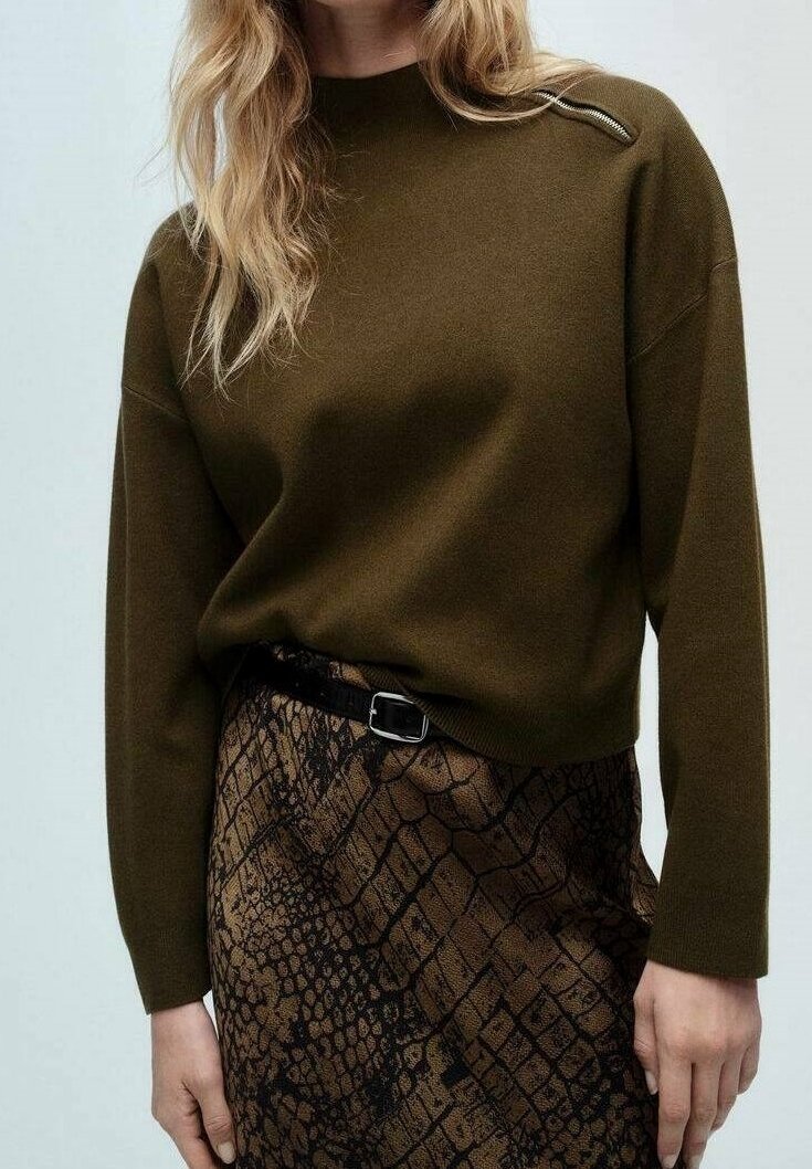 Femme portant un pull vert olive avec un détail de fermeture éclair sur une épaule et une jupe marron à motifs avec une ceinture noire.