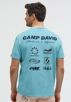 Man with dark hair wearing a light blue Camp David T-shirt with sailing and surfing themed text and graphics on the back.