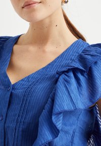 Niebieska, fakturowana bluzka z falbankami na rękawach, dekoltem w serek i guzikami z przodu. Materiał ma subtelne pionowe paski.
