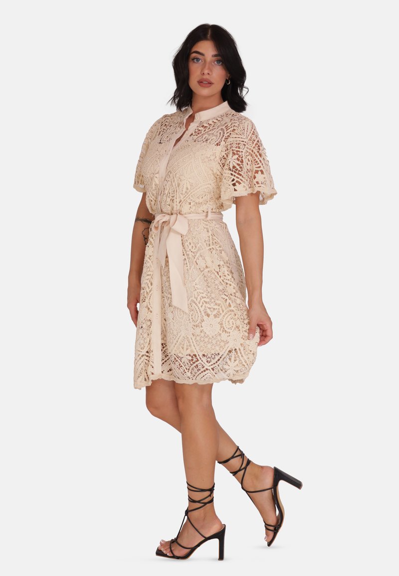Koronkowa sukienka w miękkim beżu, z kołnierzykiem, krótkimi, rozszerzającymi się rękawami, wiązaniem w talii oraz misternymi kwiatowymi wzorami. W zestawie z czarnymi sandałami na cienkich obcasach.
