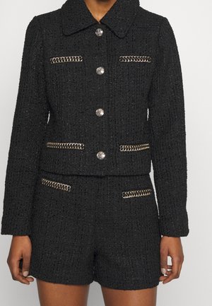 Tenue noire texturée en deux pièces avec des détails en chaîne argentée sur les poches, boutons argentés sur la veste, portée par une personne debout les bras détendus.