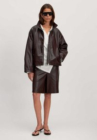 Bruine faux leren jas met rits aan de voorkant, ontspannen pasvorm, staand naast bijpassende bruine shorts. Dragen over een witte top, accessorized met zwarte zonnebril.