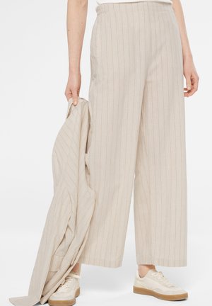 Person wearing beige pinstriped wide-leg pants and cream sneakers, holding matching beige pinstriped jacket in one hand.