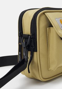 Torba Carhartt WIP z żółtego płótna o wyraźnej formie, z podwójnymi czarnymi zamkami błyskawicznymi i regulowanym czarnym paskiem. Z przodu znajduje się kieszeń z detalem.