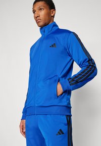 Modra poliester tracksuit jakna z visokim ovratnikom, črnimi črtami ob rokavih in Adidas logotipom na prsih.