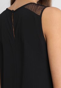 Haut sans manches noir avec un panneau transparent aux épaules et une fermeture à bouton au cou à l'arrière. Tissu doux et léger avec une texture lisse.