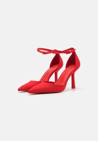 Červené lodičky na vysokom podpätku s špicatou špičkou, elegantným dizajnom a remienkom okolo členka. Hladká, matná textúra s úzkym stiletto podpätkom.