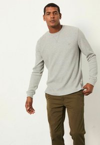 Graues Langarmshirt mit Rundhalsausschnitt, geripptem Muster und seitlichen Etiketten, kombiniert mit olivgrünen Hosen, die eine lässige Passform zeigen.