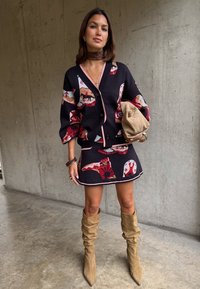 Γυναίκα με μαύρη ζακέτα με μοτίβα και φούστα, ταμπά άνετα μπότες, κρατώντας μπεζ τσαντάκι, στέκεται μπροστά σε τσιμεντένιο τοίχο.