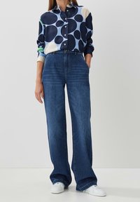 Långärmad, knäppt skjorta i blått med stora cirkulära mönster, parat med högmidjade, vida mörkblå jeans och vita sneakers.