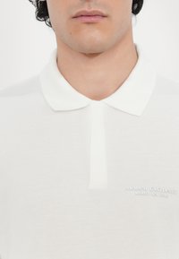 Biela polo tričko vyrobené z texturovanej tkaniny, s klasickým golierom a zapínaním na tri gombíky. Vyšívané logo "Armani Exchange".