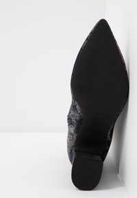 Chaussure à bout pointu fabriquée dans un tissu foncé texturé avec un motif coloré. La semelle est en caoutchouc noir avec des rainures horizontales pour une meilleure traction.