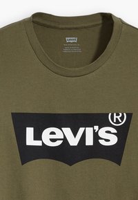 T-shirt de algodão verde-azeitona com gola redonda, apresentando um grande logótipo preto da Levi's com contorno branco e uma aba vermelha distinta na manga.