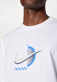 Λευκό βαμβακερό t-shirt με γραφικό σχέδιο σε μπλε και μαύρο, που περιλαμβάνει το λογότυπο της Nike, σχέδια επιμερισμού και ένα καμπύλο γεωμετρικό στοιχείο.