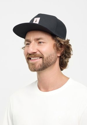 Casquette noire avec une visière plate et une pièce blanche arborant un graphisme d'arbre. Portée avec un t-shirt blanc uni. Tissu doux et design décontracté.