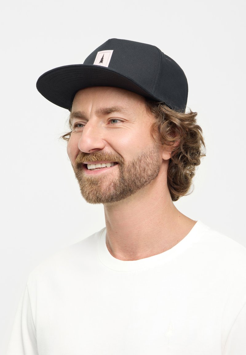 Gorra negra con un ala plana y un parche blanco con un gráfico de un árbol. Llevada con una camiseta blanca lisa. Tejido suave y diseño casual.