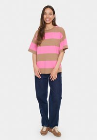 Femme souriante avec de longs cheveux bruns portant une chemise ample rayée marron et rose, un jean bleu foncé et des chaussures à lacets marron, debout.