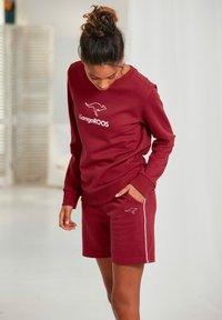 Maroon sweatshirt and shorts set with kangaroo logo. Soft fabric, relaxed fit, and white accent piping on shorts. Casual design.