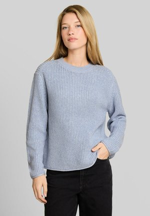 Frau mit langen blonden Haaren, die einen hellblauen Strickpullover und schwarze Hose trägt, steht mit einer Hand in der Tasche vor grauem Hintergrund.
