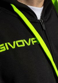 Felpa nera con vivaci stringhe verdi, logo ricamato, chiusura con zip e una trama di tessuto morbido. Ingrandimento degli elementi di design.