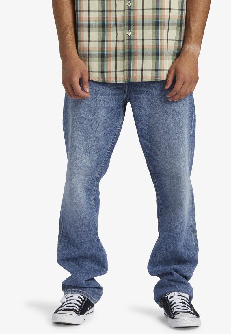 Jeans de mezclilla azul claro con corte recto, que presentan áreas desgastadas y diseño clásico de cinco bolsillos. Llevados con zapatillas negras y una camisa de cuadros.