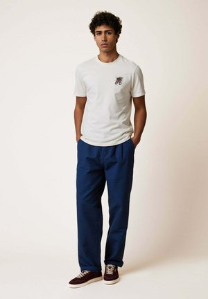 Uomo con capelli ricci che indossa una maglietta bianca con piccolo disegno e pantaloni blu, in piedi con le mani nelle tasche, sfondo neutro.
