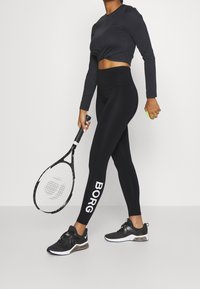 Musta pitkähihainen crop-top ja korkeavyötäröiset leggingsit, joissa on valkoinen "BORG" -logo. Henkilö pitää tennismailaa ja palloa. Mustat urheilukengät.