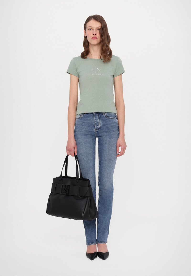 Borsa a tracolla in pelle nera con una forma strutturata e un nastro decorativo, abbinata a una maglietta verde aderente e jeans azzurri chiari.