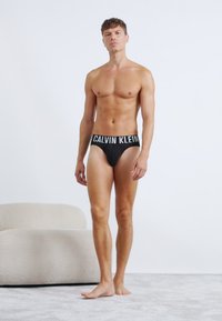 Crni Calvin Klein bokserice s širokim pojasom na kojem se nalazi bijeli natpis, prikazane na muškom tijelu protiv neutralne pozadine.