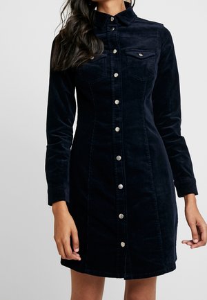 Femme portant une robe noire cintrée en velours côtelé avec des boutons, manches longues et poches poitrine, debout devant un fond uni.