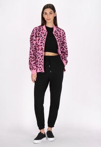 Giacca bomber con stampa leopardata rosa e polsini a coste sopra un top nero corto, abbinata a pantaloni neri a gamba affusolata e sneakers nere.