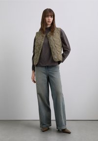 Quilted olive green vest over a dark sweater, paired with loose light blue jeans and taupe pointed-toe shoes. Smooth texture, casual design.