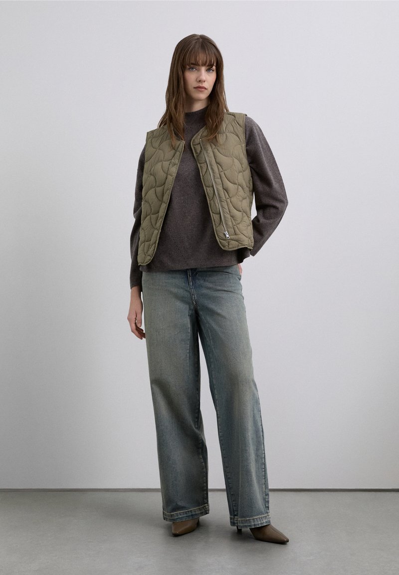 Quilted olive green vest over a dark sweater, paired with loose light blue jeans and taupe pointed-toe shoes. Smooth texture, casual design.