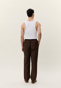 Moški stoji obrnjeno proč, oblečen v belo brezrokavnik, temno rjave hlače in rjave čevlje, ena roka je v žepu.