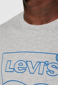 Gros plan sur une personne portant un T-shirt gris clair avec un logo Levi's à contour bleu imprimé sur le devant.