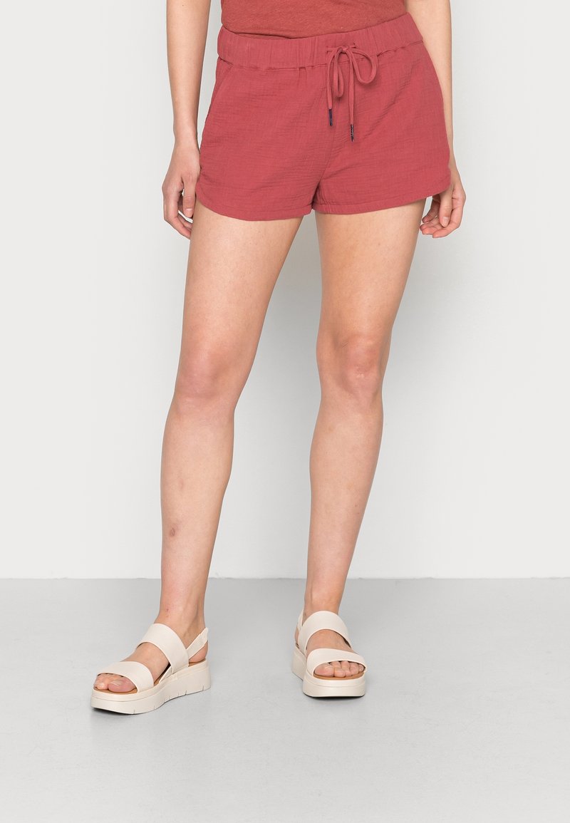 Pantalones cortos de un rojo óxido hechos de una tela texturizada, con una cinturilla elástica y cordón ajustable, y bolsillos laterales. Combinados con sandalias de plataforma.