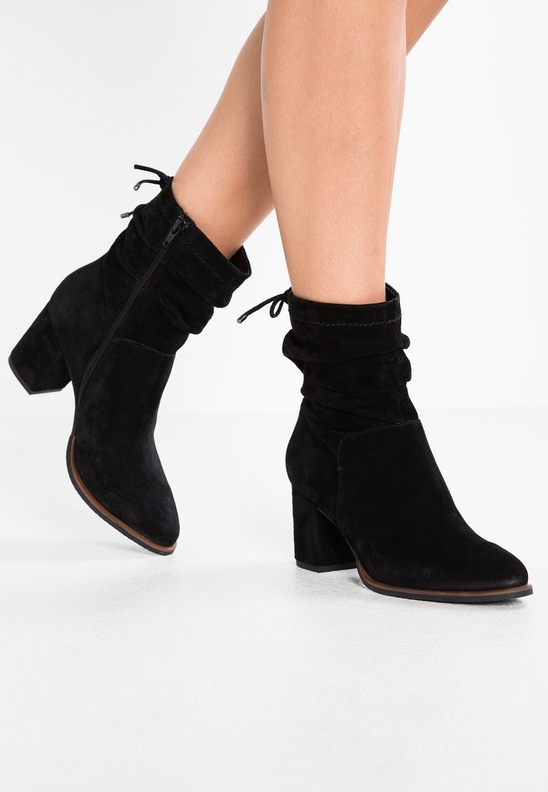 Bottines en daim noir avec un design ample, une fermeture éclair, et un talon carré. Texture lisse avec un bout arrondi et des accents en dentelle.