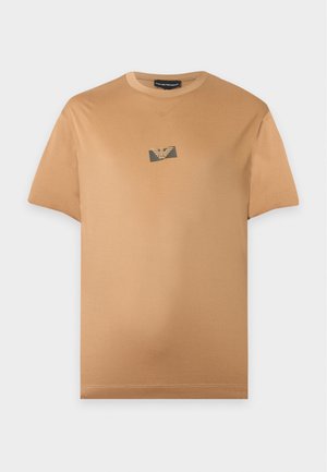 Bărbat purtând un tricou maro deschis cu mâneci scurte, blugi albaștri și adidași albi, stând în fața unui fundal simplu și deschis la culoare.