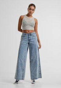 Stříbrný flitrový crop top ve střihu tank top, spárovaný s vysokým pasem a volnými světle modrými džíny. Modelka má na sobě stříbrné špičaté kotníkové boty.