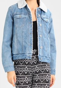 Giacca di jeans azzurro chiaro con colletto bianco e peloso, tasche frontali e bottoni metallici, abbinata a pantaloni a fantasia bianco e nero.