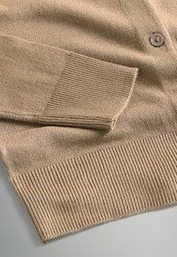 Beżowy sweter z dzianiny z rękawami zakończonymi ściągaczem, ściągaczem u dołu oraz jednym beżowym guzikiem, leżący na płaskiej powierzchni.